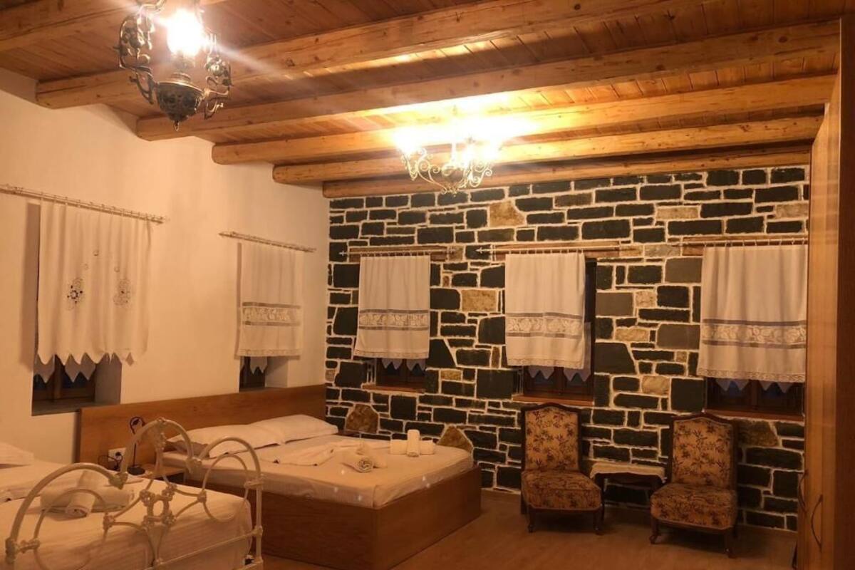 A room with a stone wall, wooden ceiling beams, a bed with a metal frame, two upholstered chairs, and a chandelier.