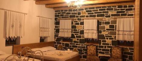 Quarto com parede de pedra, vigas de madeira no teto, cama com estrutura de metal, duas poltronas estofadas e um candelabro.