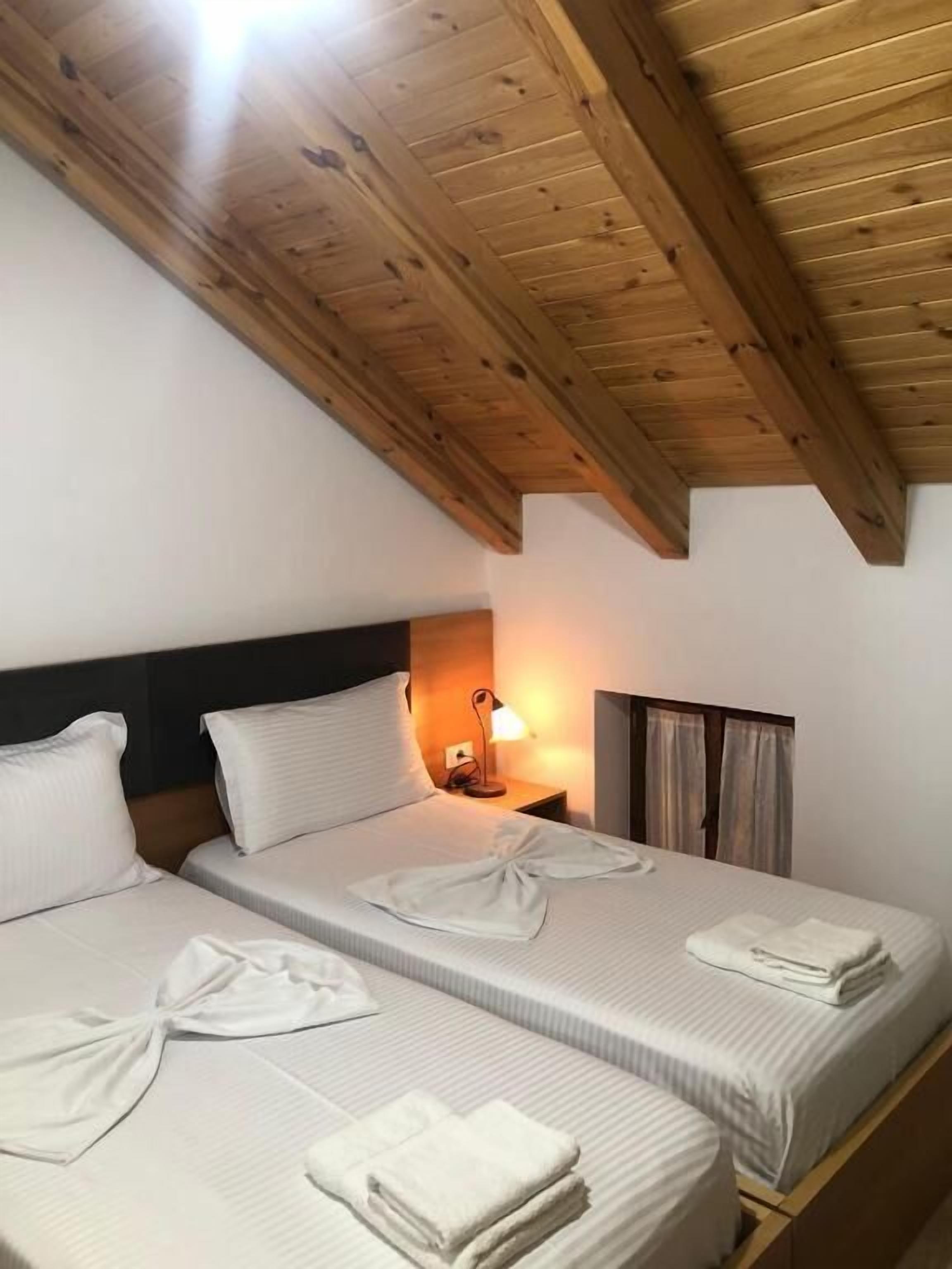 A room with two beds, wooden ceiling beams, a nightstand with a lamp, and a window with curtains.