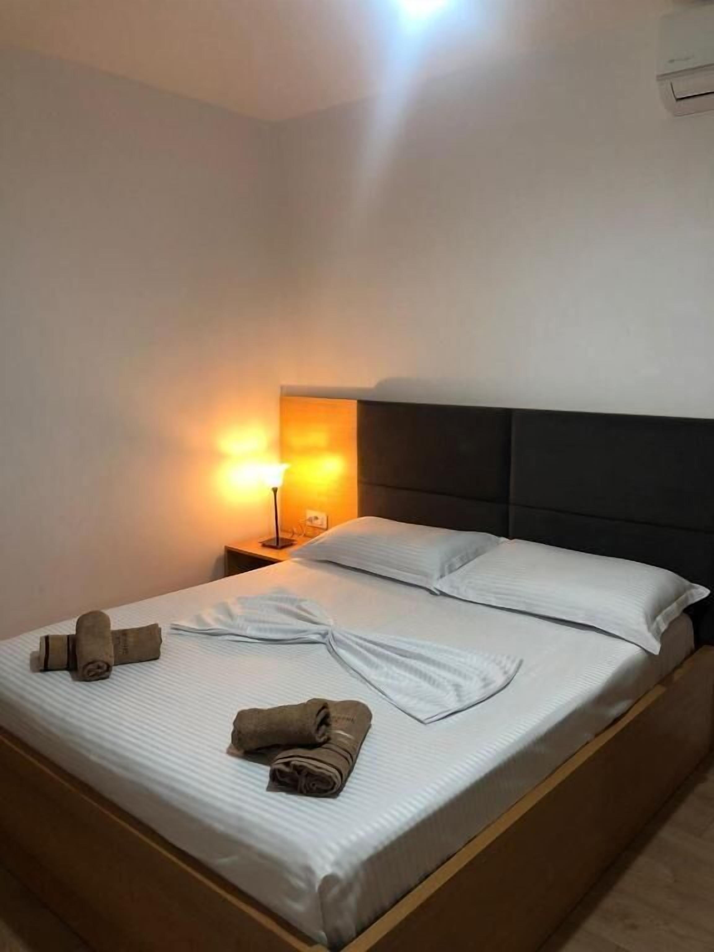 Uma cama bem arrumada com lençóis brancos, duas toalhas enroladas e uma luminária de cabeceira.