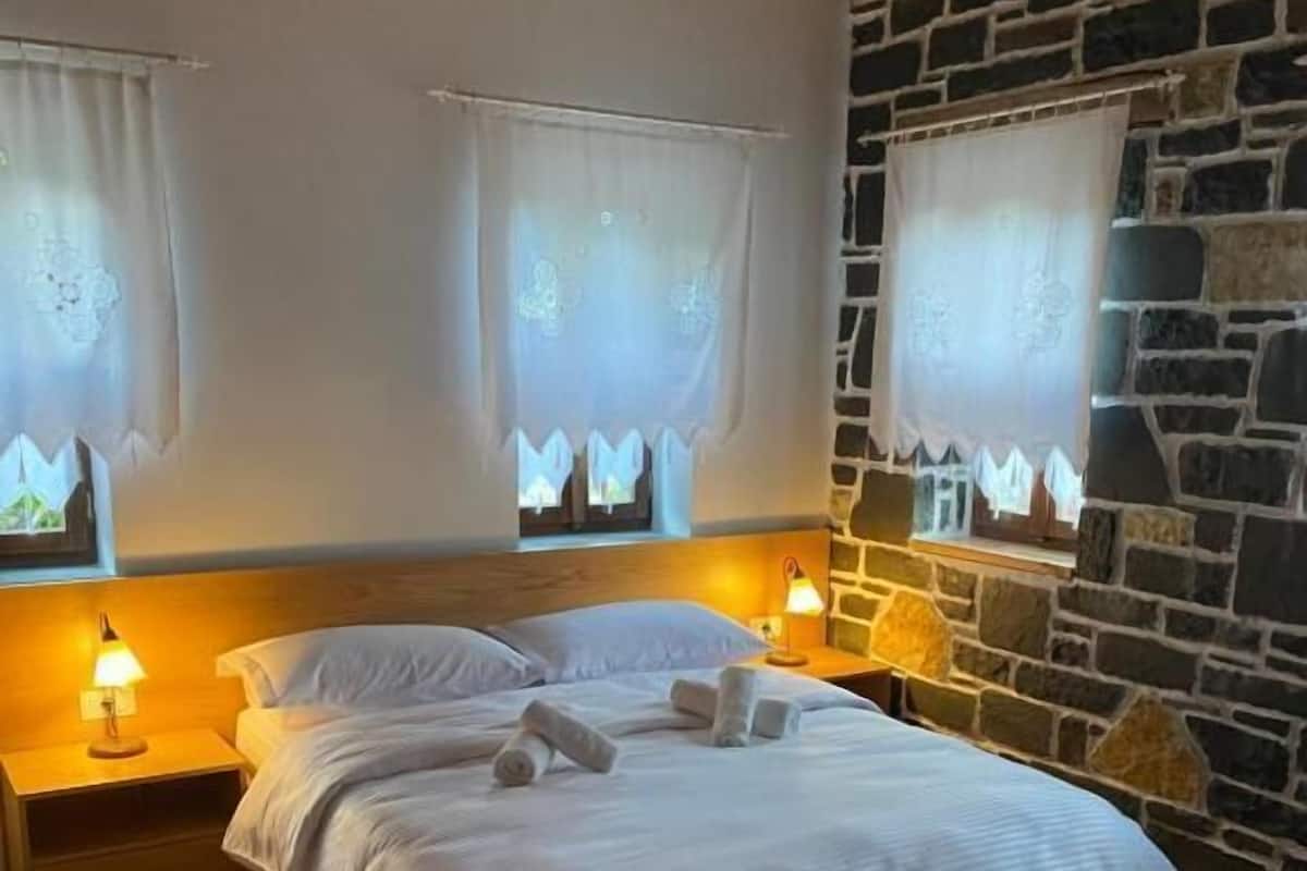 A bedroom with a wooden headboard, a large bed with white linens, two bedside lamps, and a stone wall.