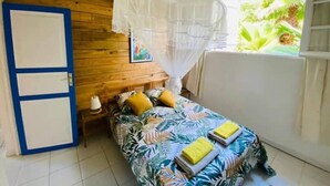 A room with a bed, a nightstand, a blue door, and a window with a view of greenery. - La Casa Colibris (Anse-Bertrand)