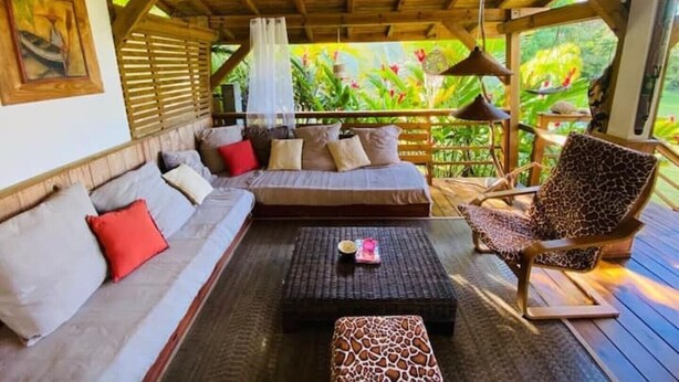 A covered outdoor seating area with a sofa, a patterned ottoman, and a chair. - La Casa Colibris (Anse-Bertrand)