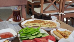 Een tafel met diverse gerechten, waaronder een bord pannenkoeken, een bord brood, een bord gesneden komkommers, tomaten en groene paprika's, een bord croissants, een bord chocoladecake en een glas thee.