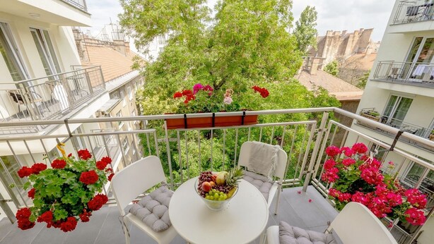 Ein Balkon mit rundem Tisch, zwei Stühlen und einer hängenden Pflanzampel mit roten Blüten.