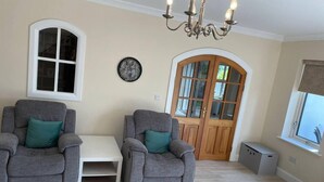 A living room with two grey armchairs, a white coffee table, a chandelier, a wooden door, and a window with blinds. - Beautiful Home Near Lough Corrib (Carrowmoreknock)