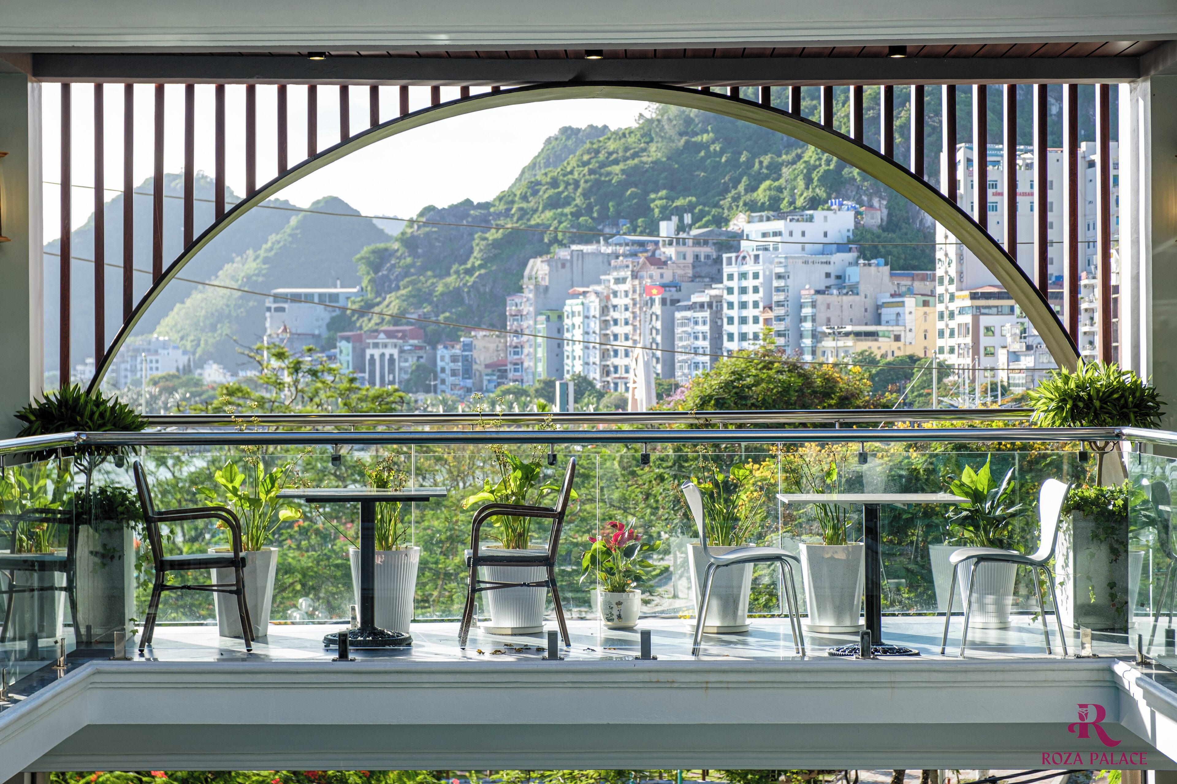 A balcony with a glass railing, potted plants, and a view of a cityscape with buildings and greenery.