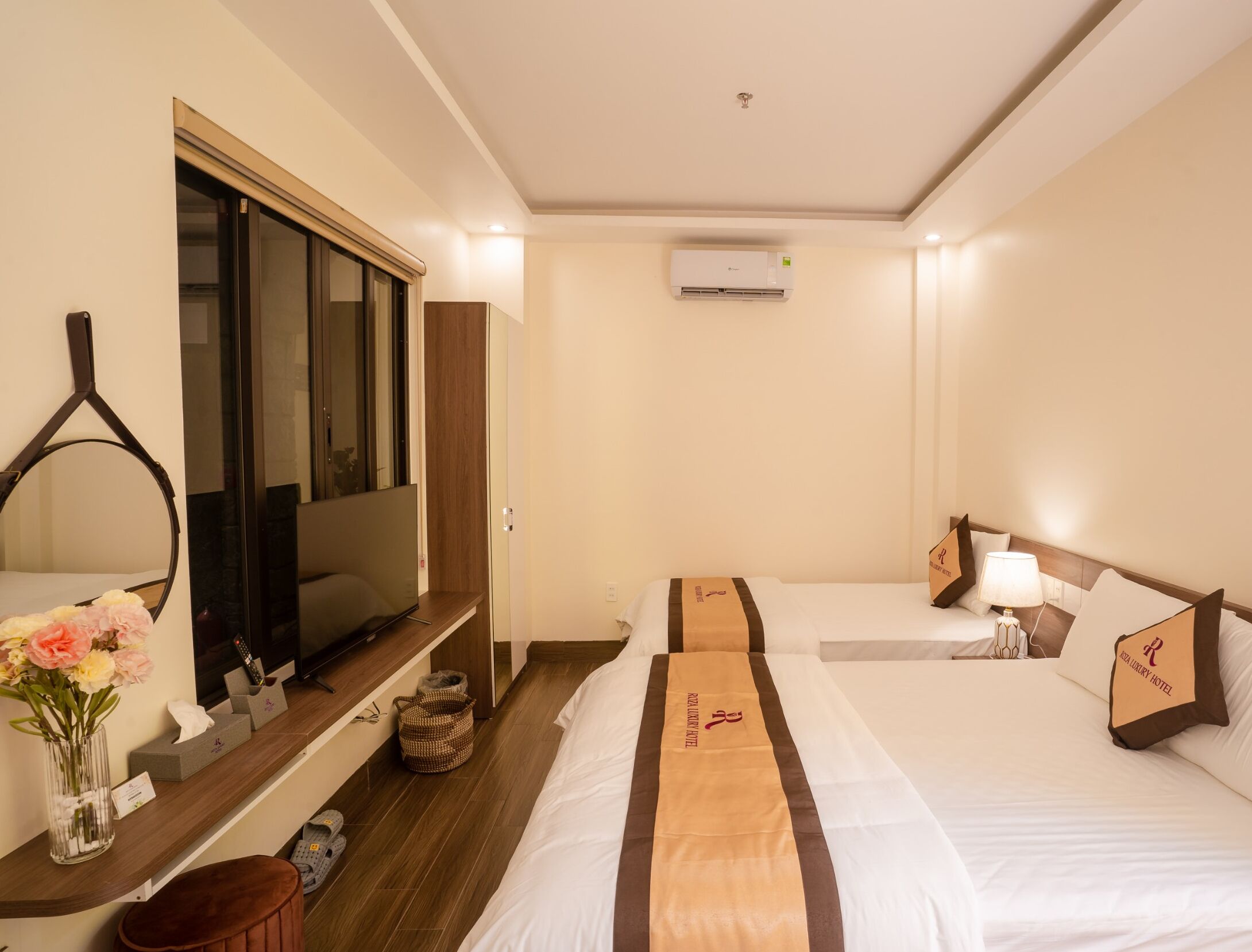 A hotel room with a TV, a bed with a brown headboard, a bedside table with a vase of flowers, and a wooden floor.