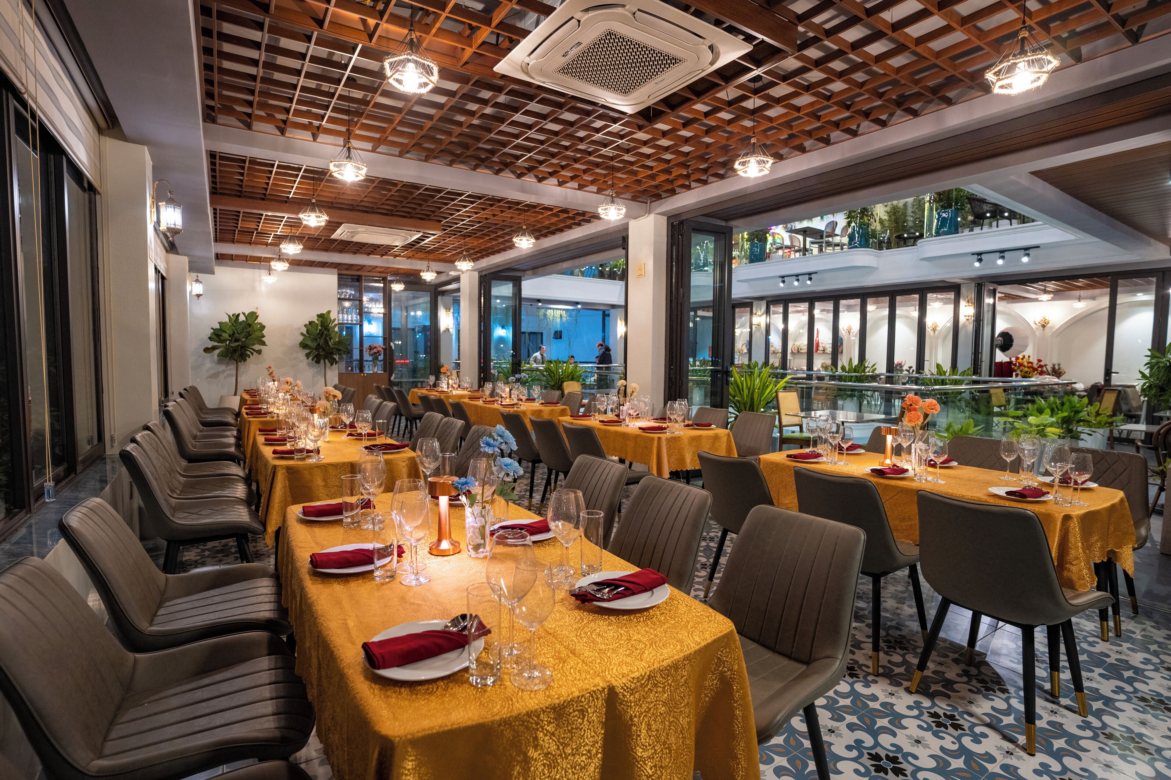 A restaurant with long tables set for dining, featuring a modern interior with wooden ceiling beams and large windows.