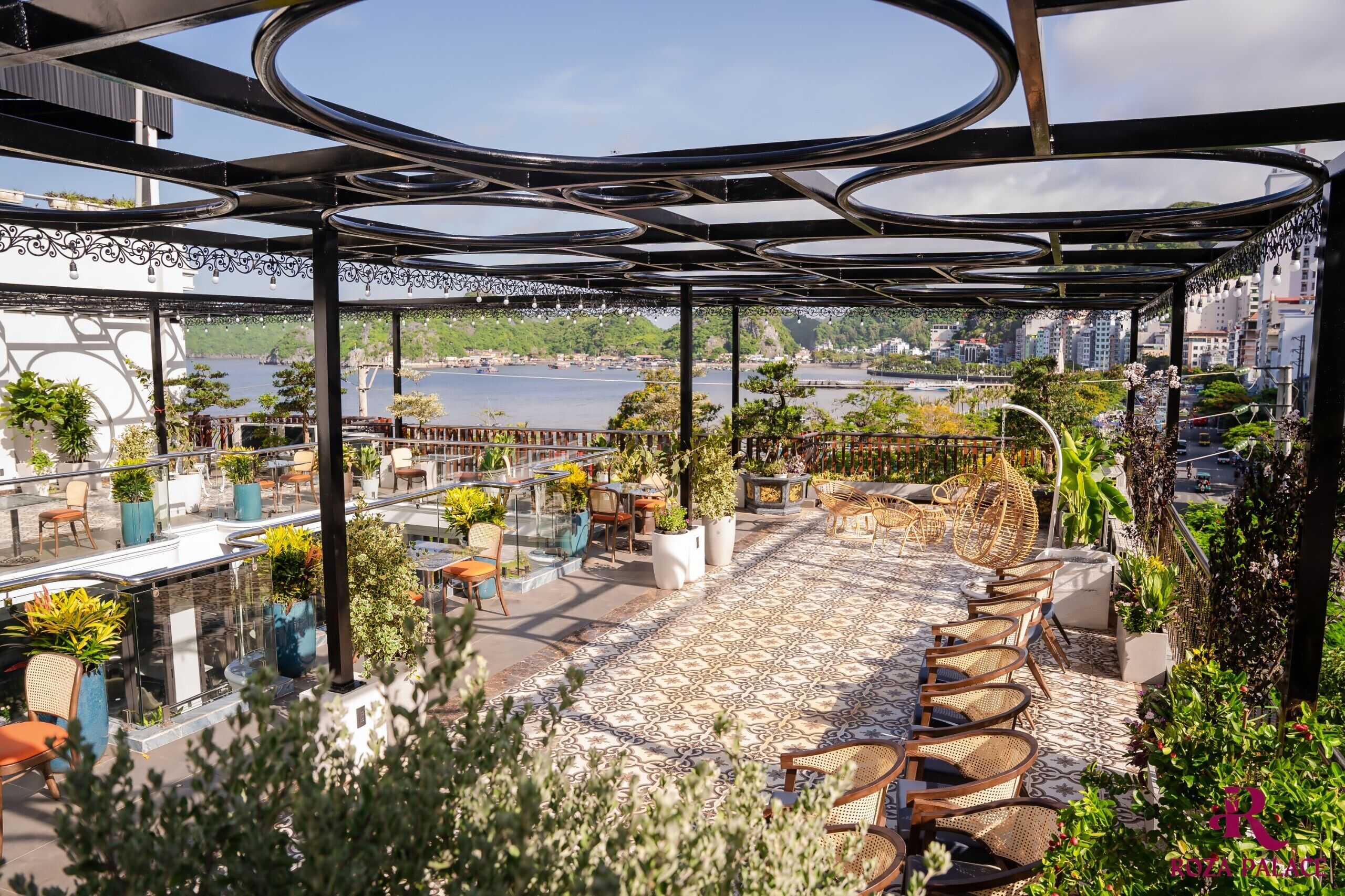 An outdoor terrace with a view of a body of water, furnished with chairs and tables, surrounded by greenery.