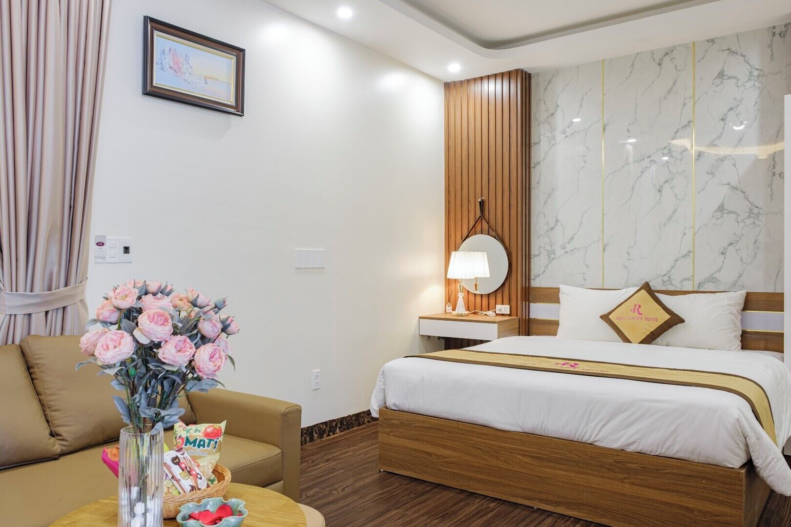 A hotel room with a bed, a wooden nightstand, a lamp, a vase with flowers, a framed picture, and a wall with a marble-like pattern.
