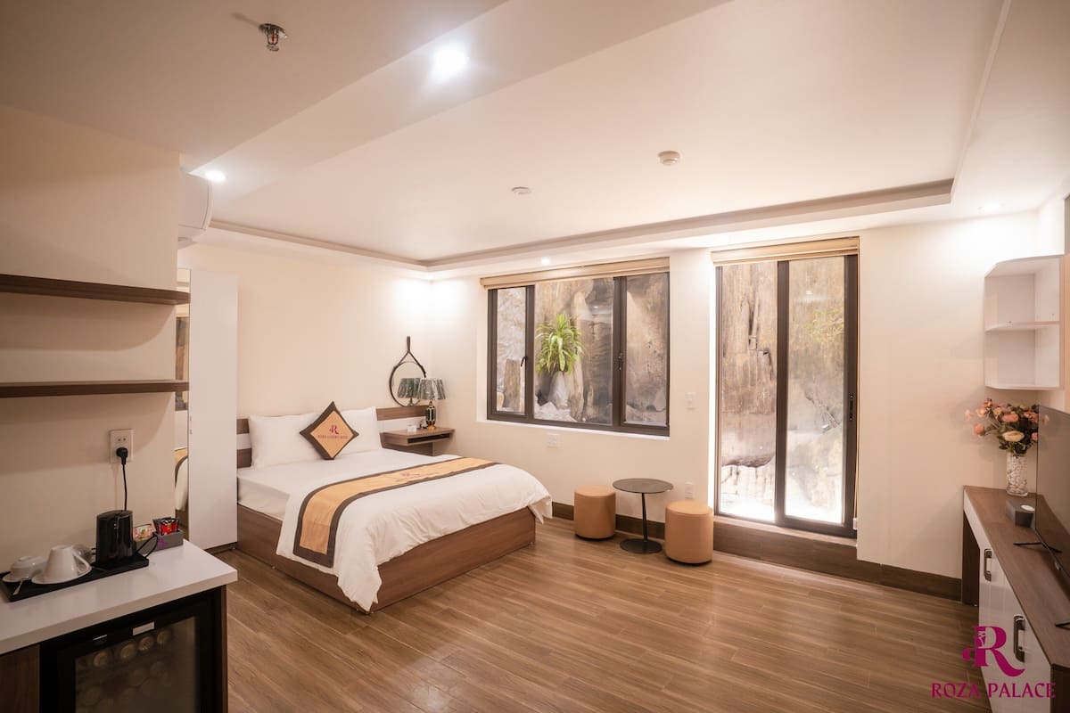 Una habitación de hotel moderna con una cama grande, una mesita de noche con lámpara, una mesa redonda pequeña y un amplio ventanal con vistas a un paisaje rocoso.