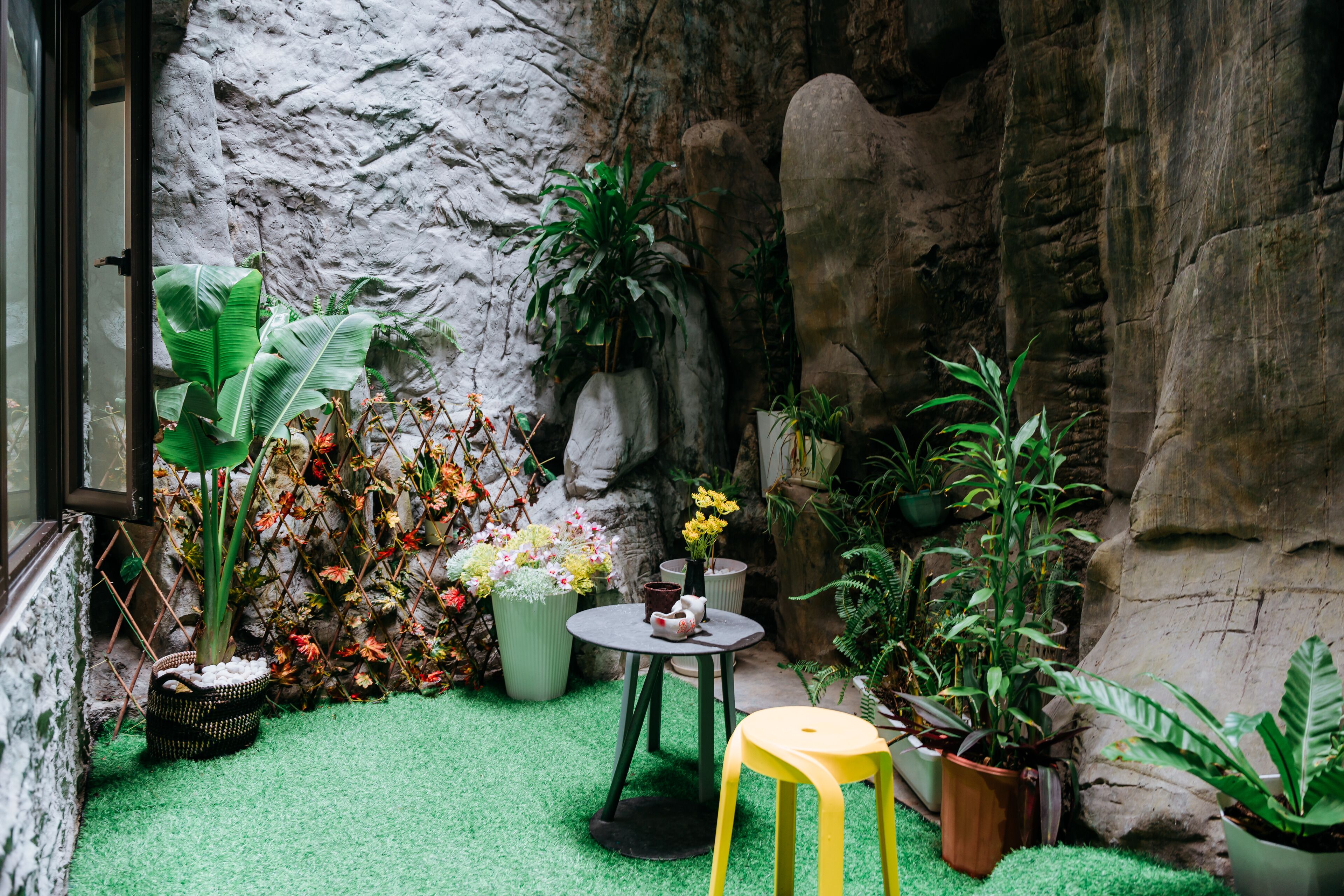 A small, enclosed garden area with artificial grass, potted plants, a small table, and a yellow stool.