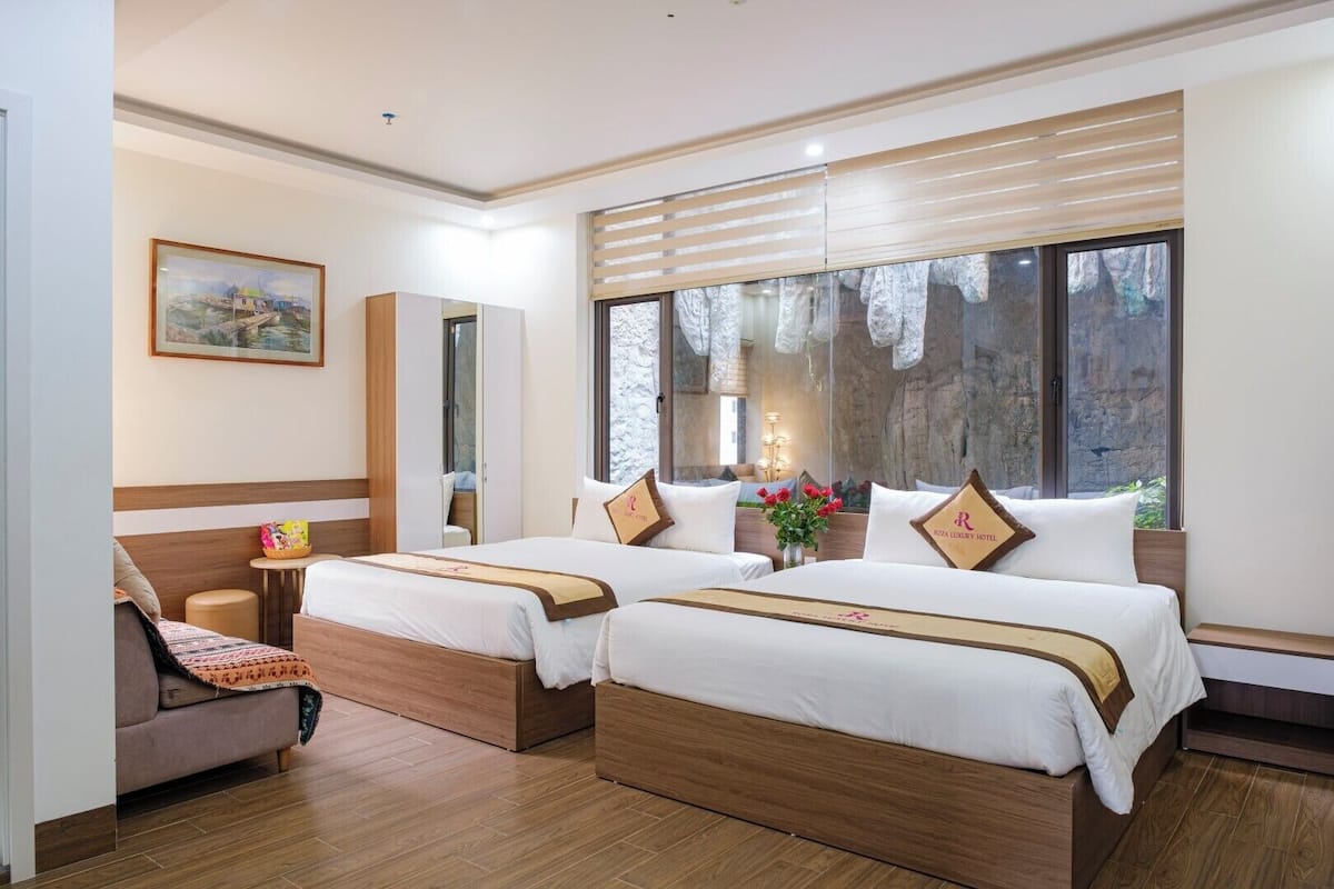Habitación de hotel con dos camas, un sofá, una mesita, un cuadro en la pared y un ventanal con vistas a árboles.