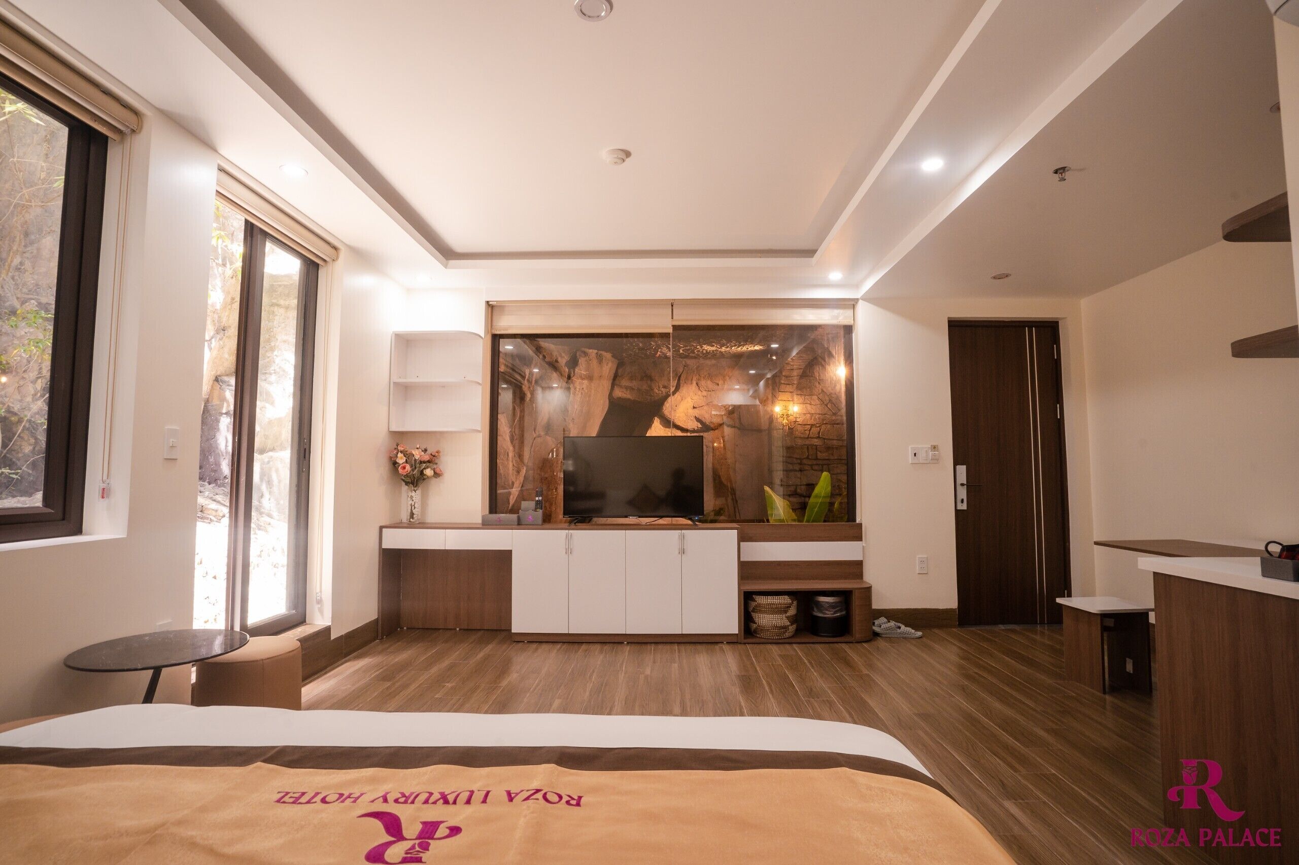 Una habitación de hotel moderna con una cama grande, un televisor, un mueble de madera, una mesita pequeña y un ventanal amplio.