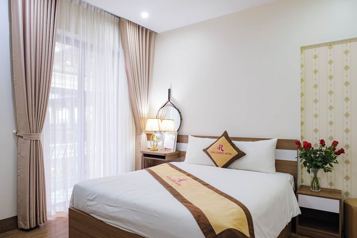 Habitación de hotel con una cama grande, una mesita de noche con lámpara, un jarrón con flores y una ventana con cortinas.