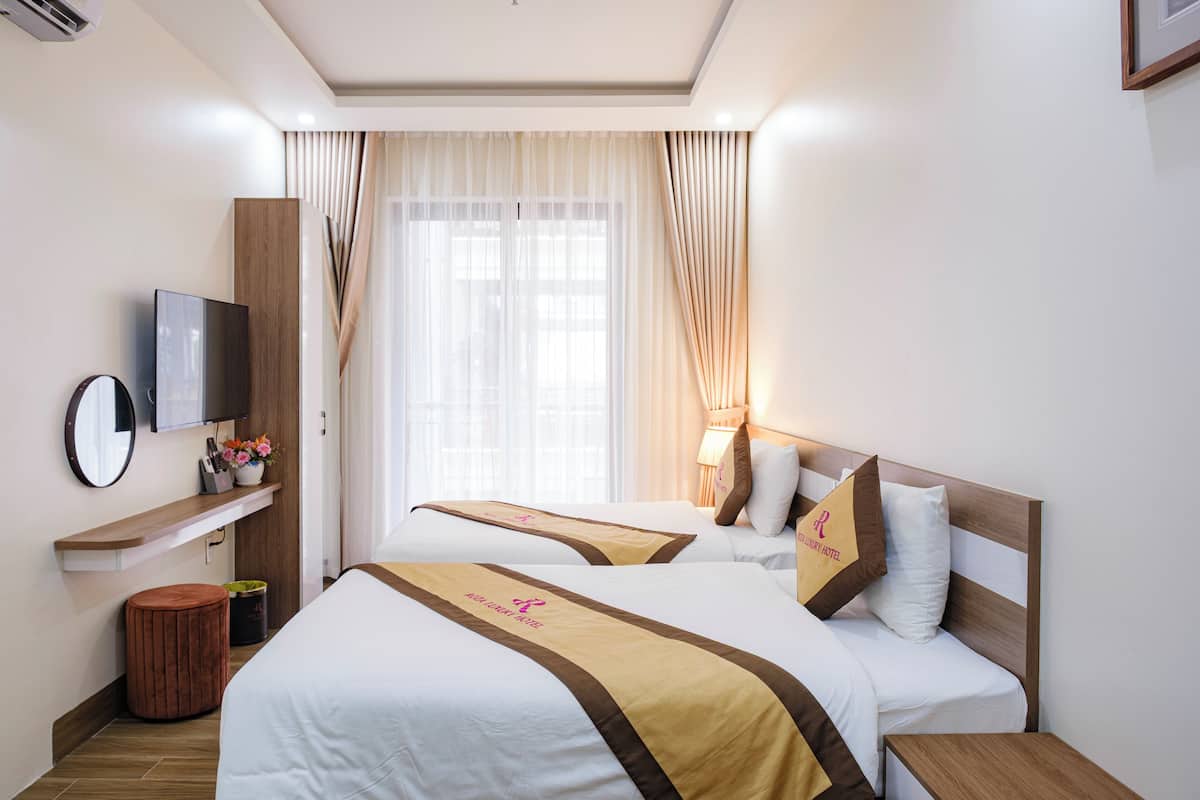 Habitación de hotel con dos camas, un televisor, un estante de madera con un jarrón y una ventana con cortinas.
