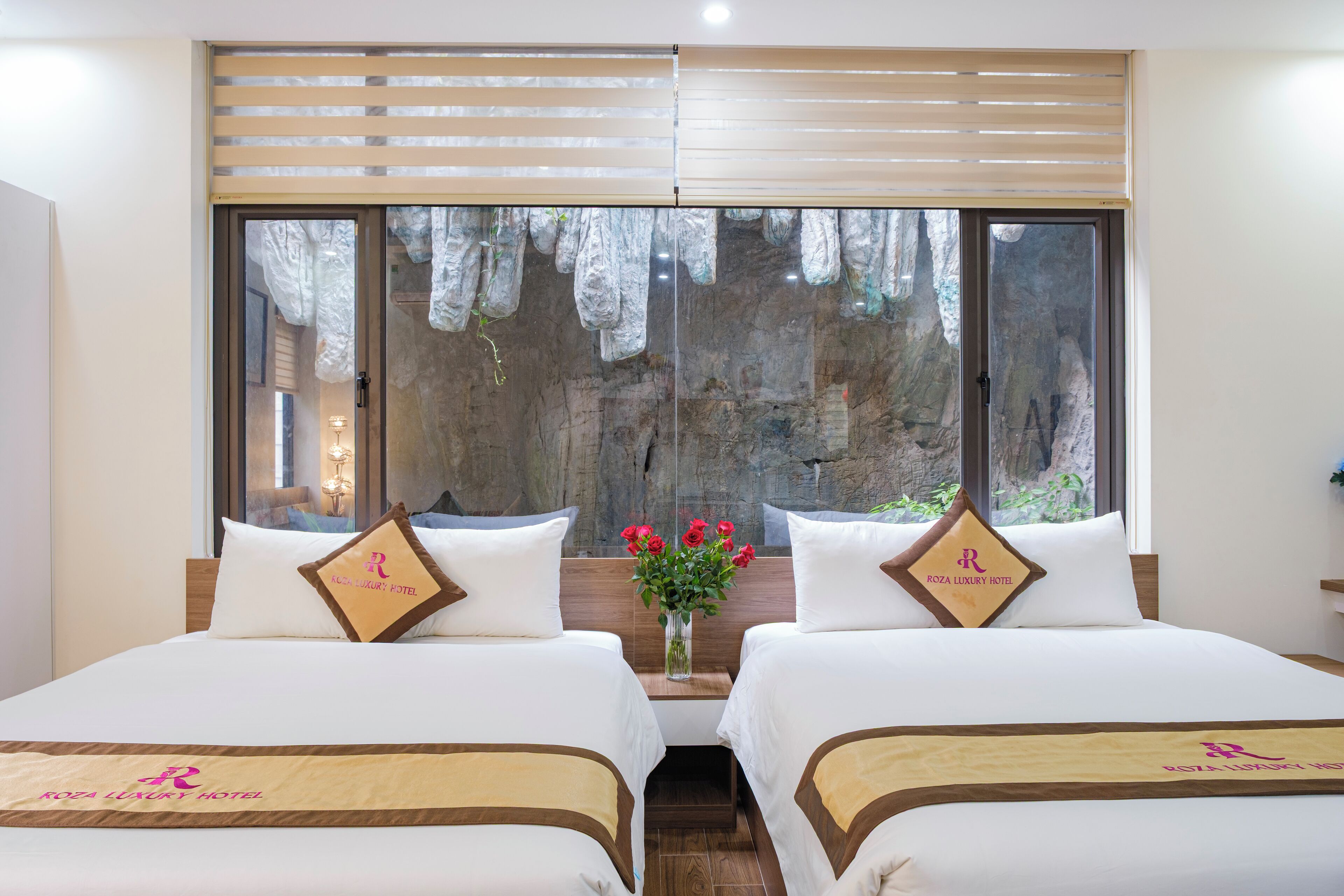 Una cama doble con cabecera de madera, una ventana con vistas a un paisaje rocoso y un florero con flores rojas en la mesita de noche.