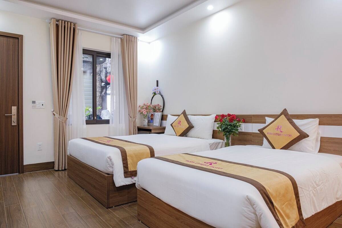 Una habitación de hotel con dos camas, cabeceros de madera, cortinas color beige, un ventanal con vistas y una mesita con un florero.