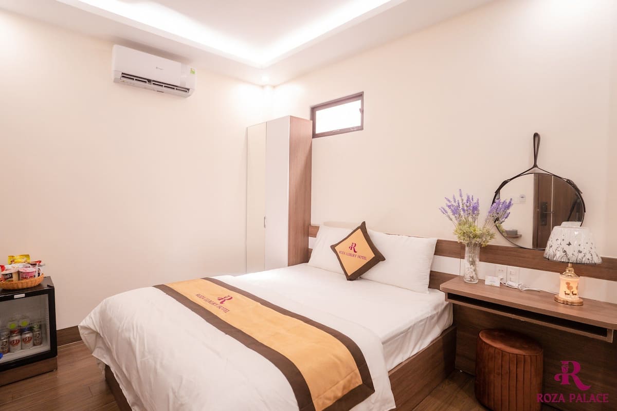 Una habitación de hotel con cama, mesita de noche, lámpara, espejo y una ventana pequeña.