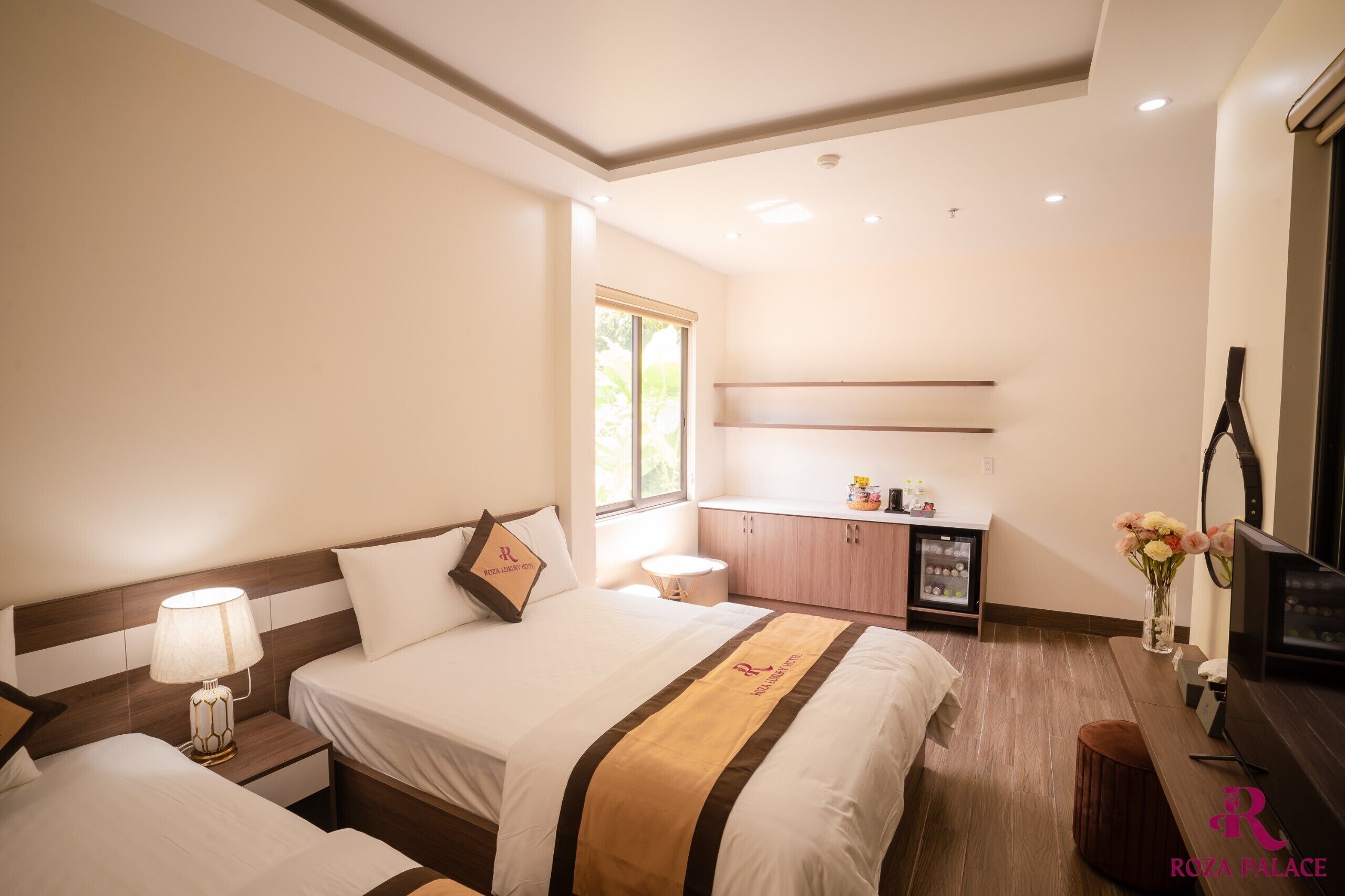 A hotel room with two beds, a desk, a TV, and a window.