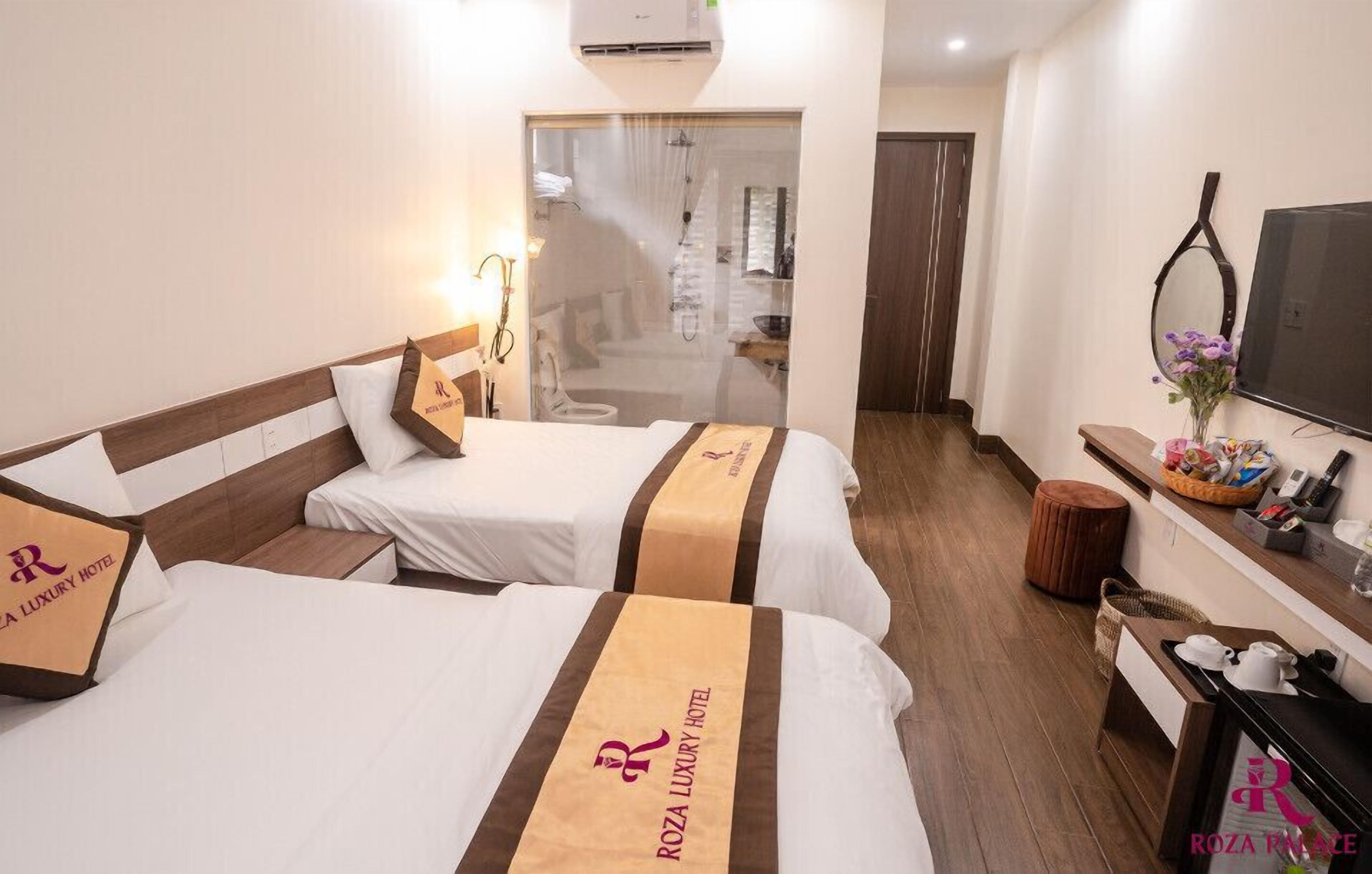 A hotel room with two beds, a desk, a TV, and a bathroom visible through a glass door.