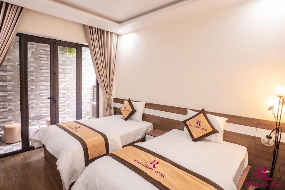Habitación de hotel con dos camas, un ventanal grande con cortinas, una mesita de noche y una lámpara.