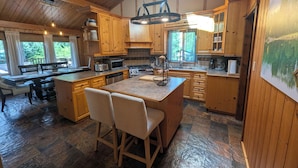 A spacious kitchen with wooden cabinets, a large island with bar stools, a dining area with a bench, and a view of the outdoors through large windows.