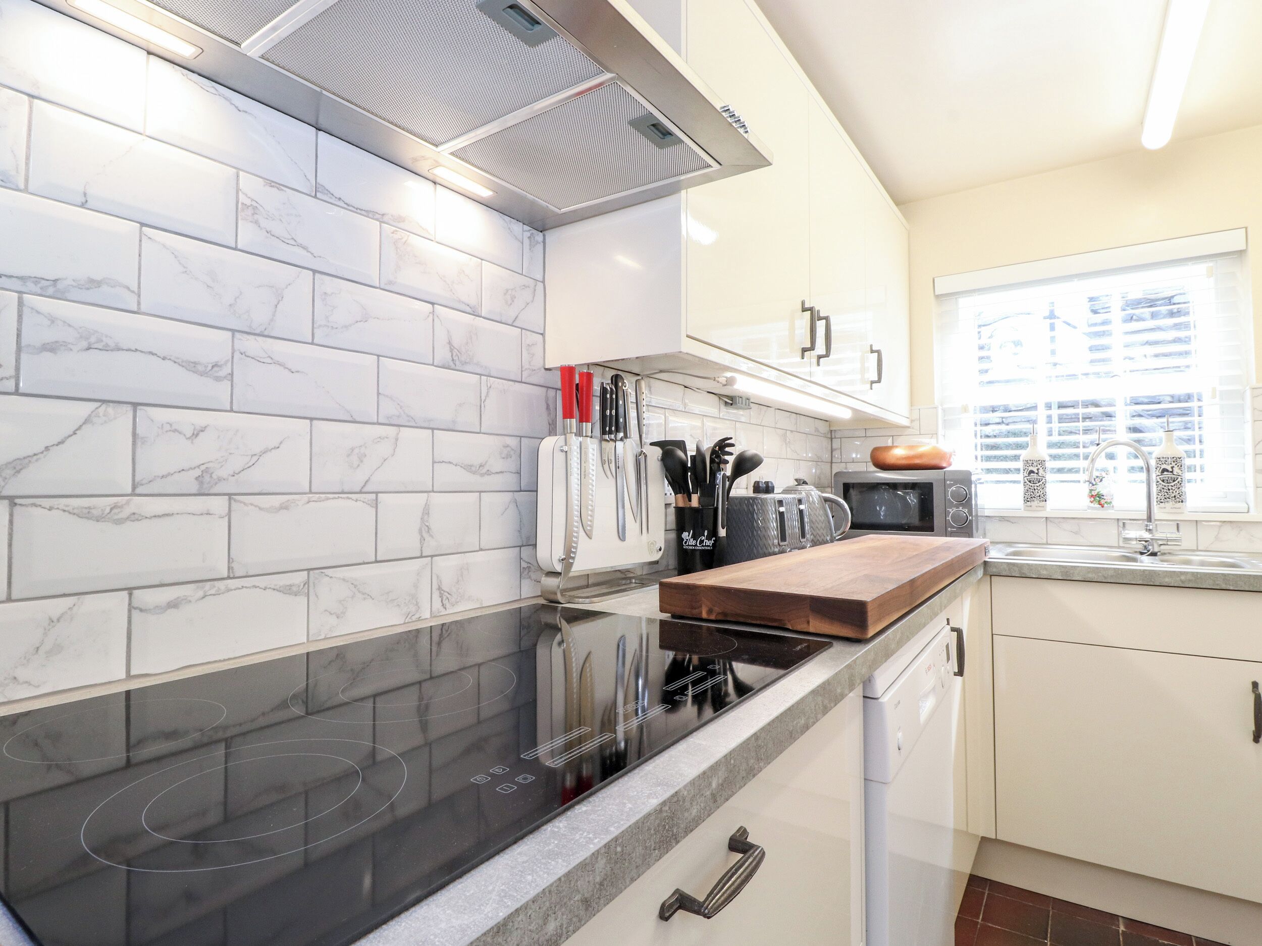 A modern kitchen with white marble tiles, a black induction cooktop, a wooden countertop, and a window with blinds.