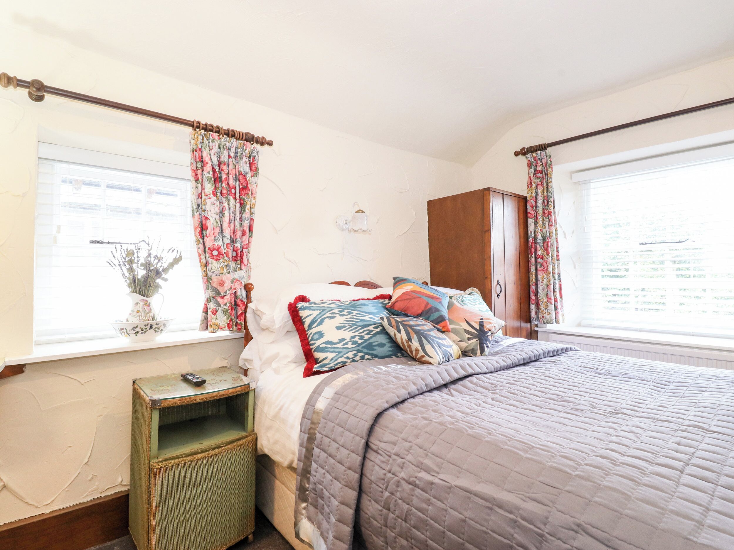 Un dormitorio con cama, cortinas florales, un armario de madera y una mesita con un florero.