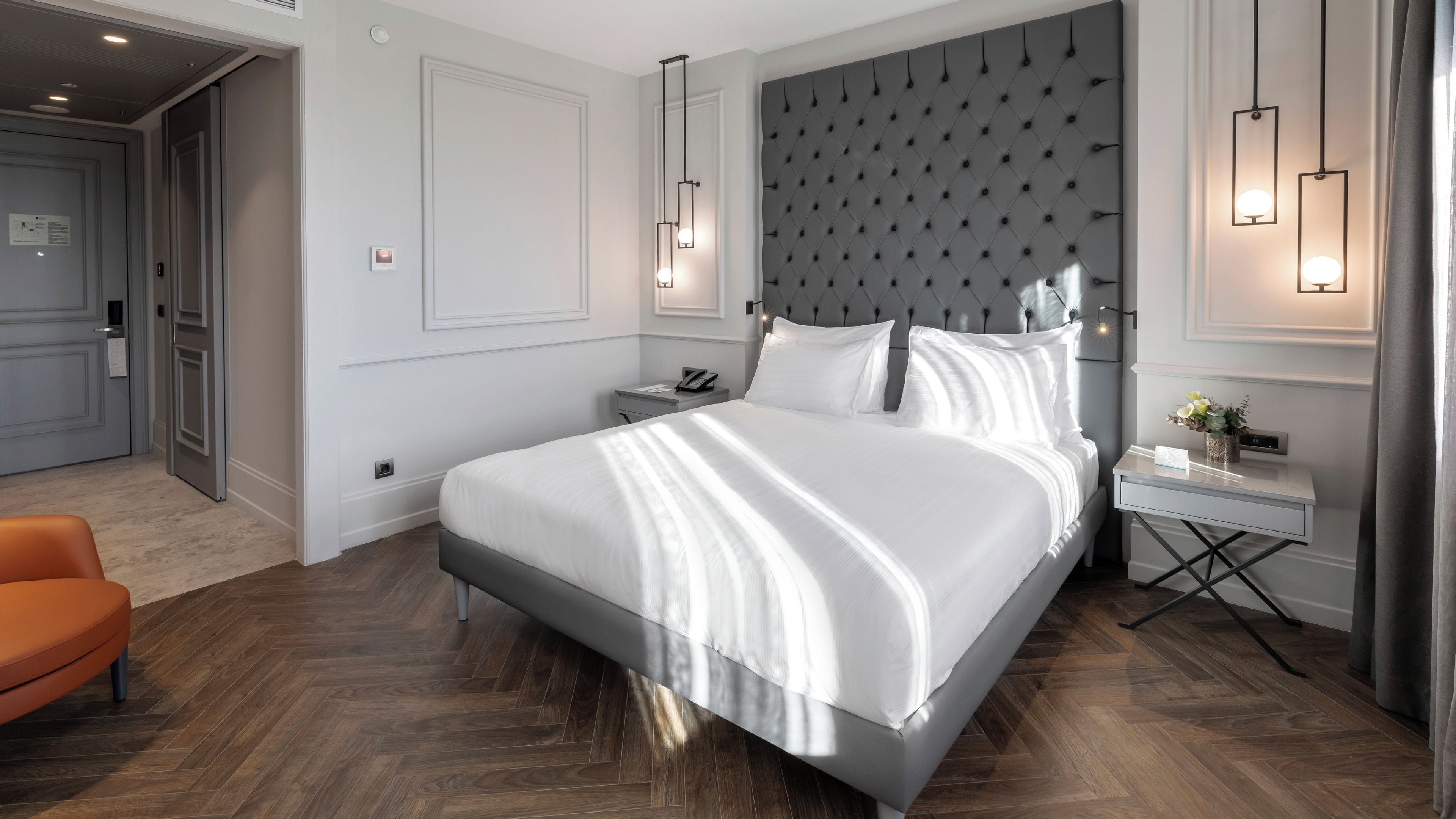 A hotel room with a large bed, a bedside table, a nightstand, a chair, and a wall-mounted light fixture.