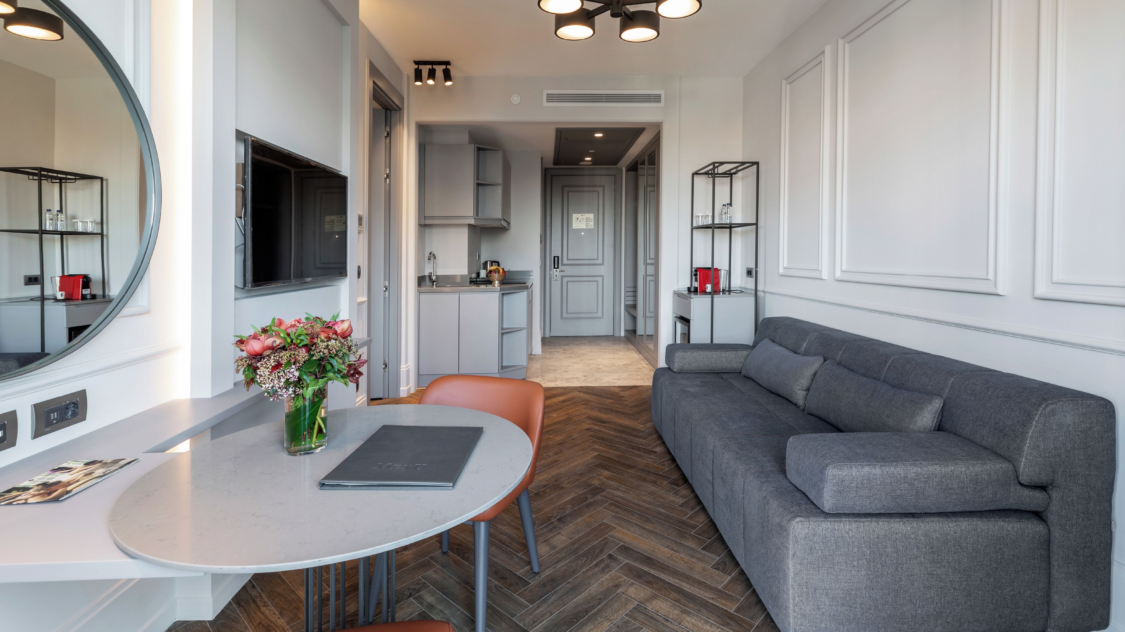 A modern living room with a grey sofa, a dining table with a vase of flowers, and a TV mounted on the wall.