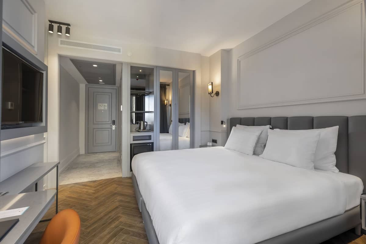 A modern hotel room with a large bed, a desk, and a chair. There is a wardrobe and a mirror in the background.