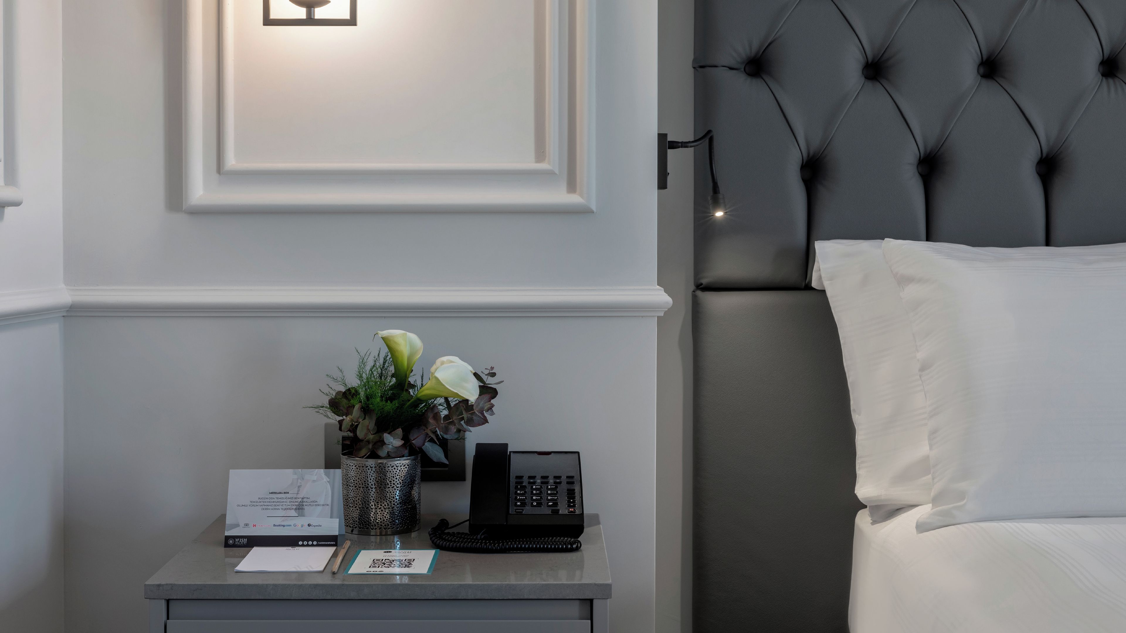 A hotel room with a neatly made bed, a bedside table with a telephone, a vase with flowers, and a framed picture on the wall.