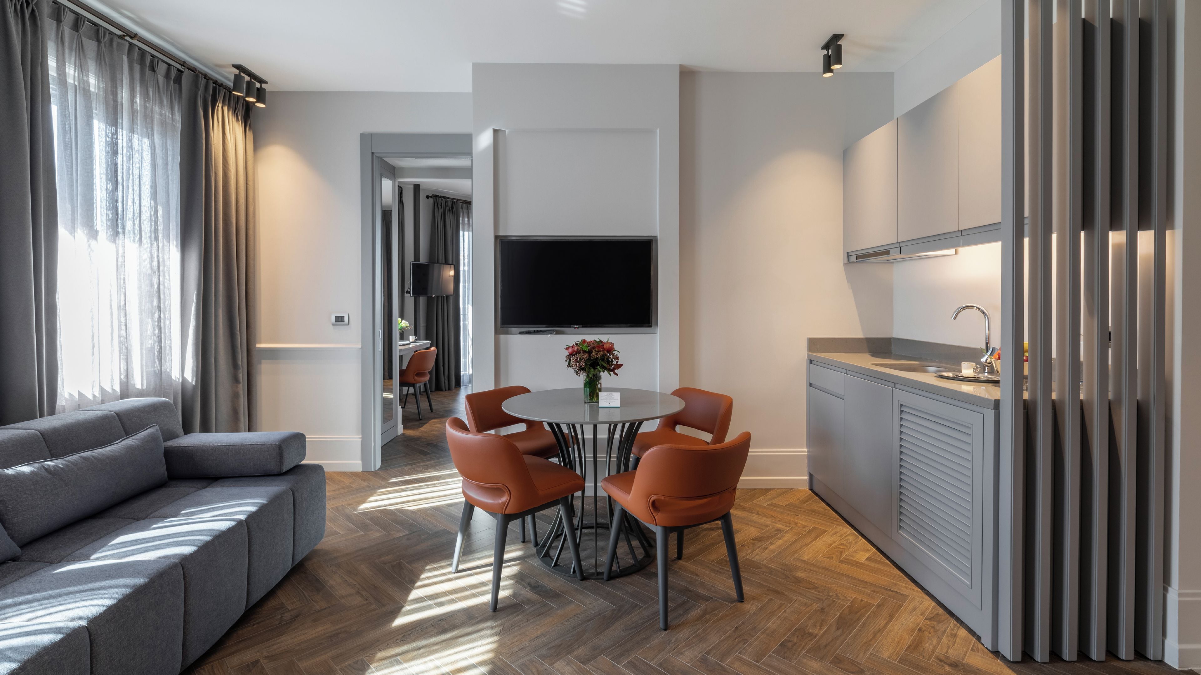 Un salon moderne avec un canapé gris, une table à manger entourée de chaises orange, un téléviseur à écran plat fixé au mur et un coin cuisine équipé d’un évier et de placards.