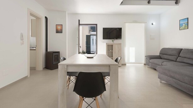 Ein modernes Wohnzimmer mit grauem Sofa, einem Esstisch mit Stühlen, einem an der Wand montierten Fernseher und einer Treppe, die in eine obere Ebene führt.