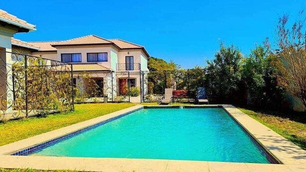 A rectangular swimming pool with a tiled edge, surrounded by a grassy area and a house with a tiled roof and a balcony. - 24 Roma (Lusaka)