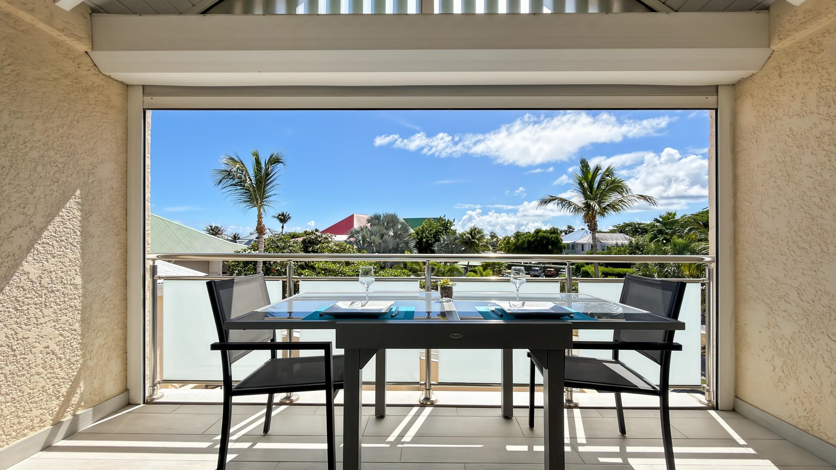 A balcony with a glass table and chairs, overlooking a tropical landscape with palm trees and a clear sky.