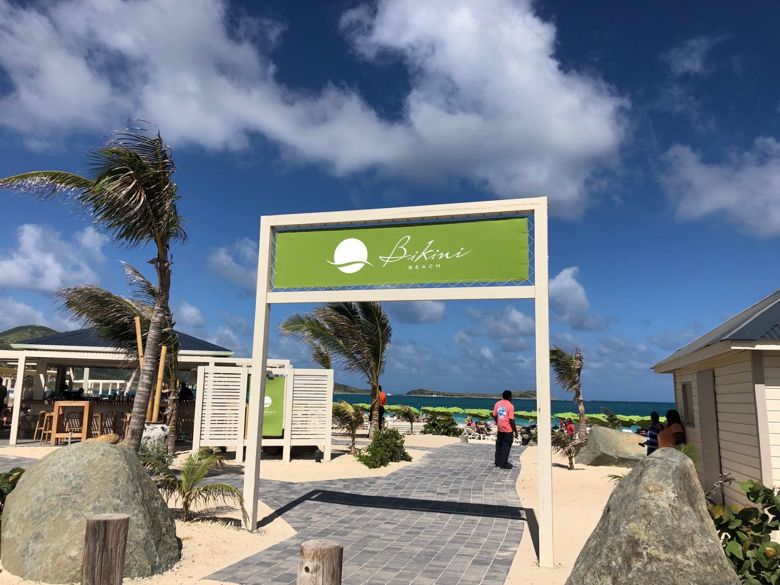 A beachside sign for Bikini Beach with a clear view of the ocean and palm trees.