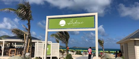 A beachside sign for Bikini Beach with a clear view of the ocean and palm trees.