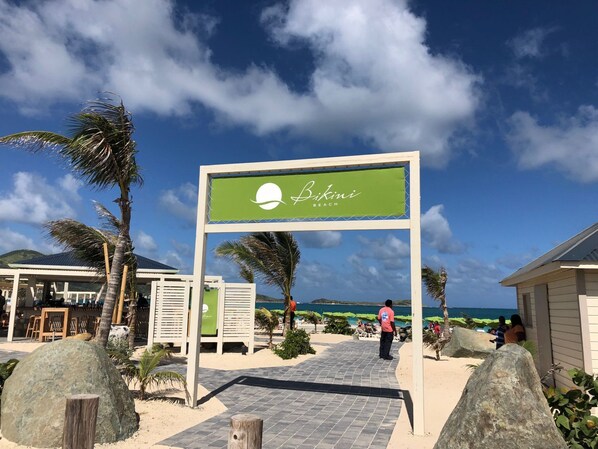 A beachside sign for Bikini Beach with a clear view of the ocean and palm trees.