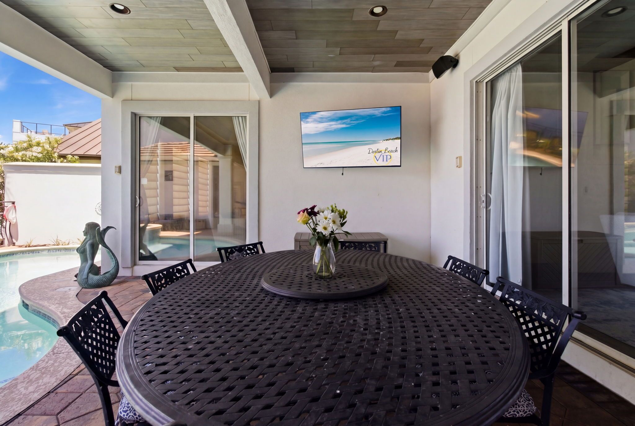 Un área de comedor al aire libre techada con una mesa redonda, sillas, un florero con flores, una piscina y un televisor con temática playera.