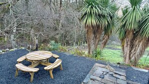 A wooden table and benches set on a gravel area with a stone border, surrounded by trees and a fence. - The Old Schoolhouse - Character Stay on NC500 (Tongue)