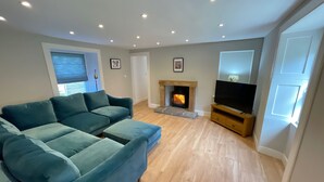 A living room with a sectional sofa, a fireplace, a television on a wooden stand, and a door with a window. - The Old Schoolhouse - Character Stay on NC500 (Tongue)