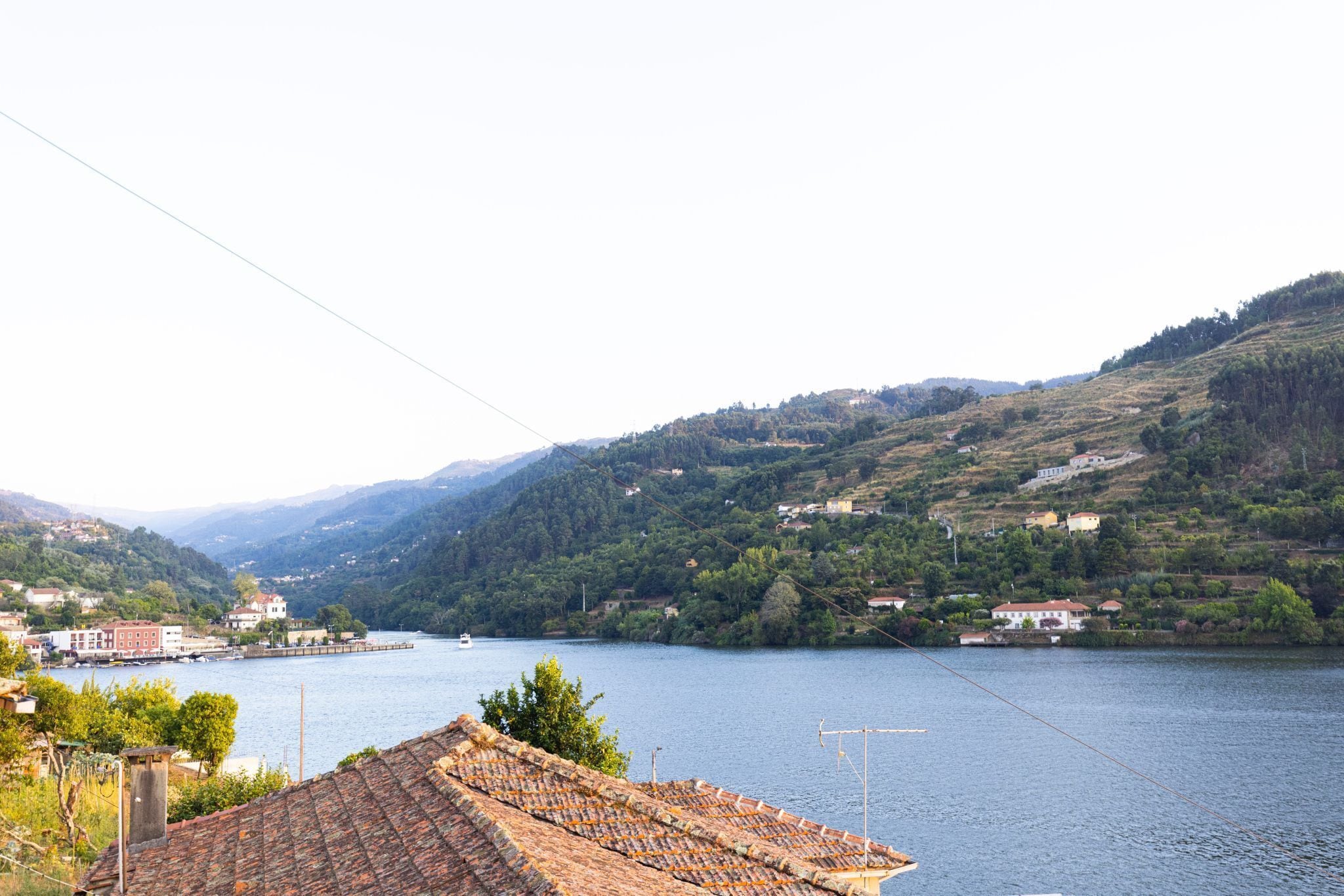 A riverside view with terracotta-roofed buildings, a dock, and hills in the background.