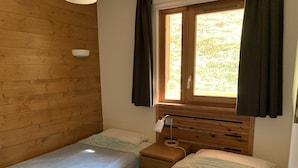 Een kleine, knusse kamer met twee eenpersoonsbedden, een houten hoofdbord, een nachtkastje met lamp en een raam met gordijnen.
