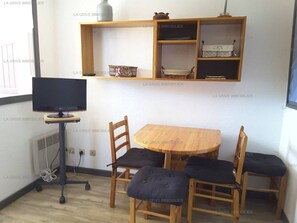 Un petit coin repas avec une table ronde en bois et quatre chaises, un téléviseur posé sur un meuble, et une étagère au-dessus de la table où sont rangés divers objets.