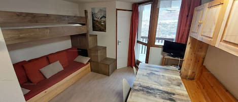 Ein kleines Zimmer mit einem Etagenbett, einem roten Schlafsofa, einem kleinen Tisch und einem Fernseher.