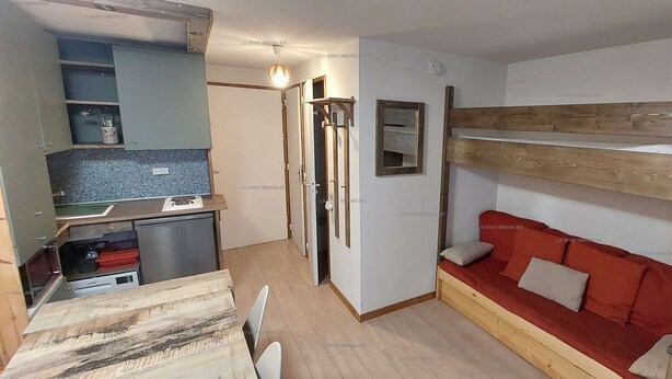 A compact room with a kitchenette, a bunk bed with red bedding, and a small seating area.