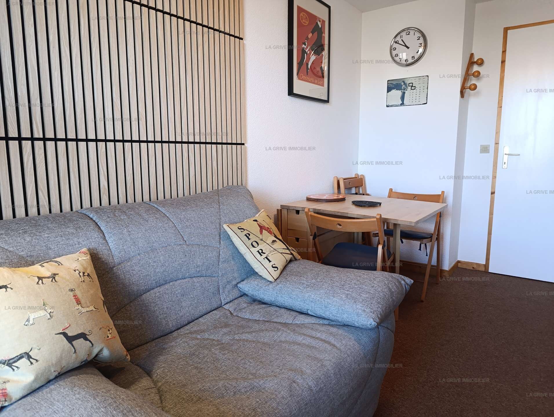 A living room with a grey sofa, a small table, and a chair. There is a clock and a framed picture on the wall.